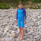A boy stands on a pebbled beach in a 'UV Protective Rash Vest Blue Starfish', smiling at the camera.